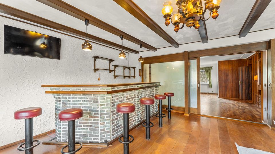 Innenraum mit gemauerter Theke im Barstil, Holzfußboden und mehreren Barhockern mit roten Polstern. Sichtbare Deckenbalken, hängende Leuchten und eine große Glasschiebetür führen in einen angrenzenden Raum mit Holzvertäfelung.