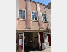 Fassade eines mehrgeschossigen Altbaus in hellem Rosa mit weißen Fensterrahmen und zwei hohen vertikalen Bannern mit der Aufschrift „MÜHLENHAUPT“ und „HÖFE“. Im Erdgeschoss führt eine breite Durchfahrt in einen Innenhof; neben dem Eingang stehen Schautafeln und Beschilderungen.