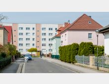 Wohnsiedlung mit einer Mischung aus Einfamilienhäusern mit roten und braunen Dächern sowie einem mehrstöckigen, weiß-rosa Mehrfamilienhaus im Hintergrund. Die Straße ist schmal, gesäumt von Gehwegen, Vorgärten und einer einzelnen, in Folie eingewickelten parkenden Stelle.