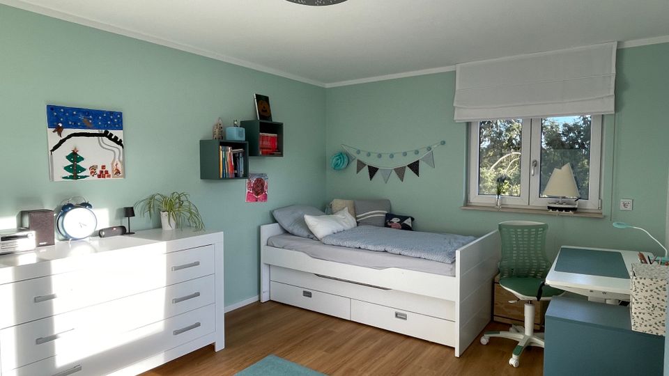 Ein Kinderzimmer mit mintgrünen Wänden, Holzfußboden und weißer Einrichtung. Zu sehen sind ein Bett mit Schubladen, ein Schreibtisch mit grünem Stuhl unter einem Fenster sowie eine Kommode mit Pflanzen und Dekoartikeln.