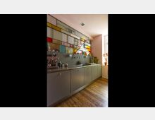 Moderne Küche mit einer Zeile aus grauen Unterschränken und einer Arbeitsfläche mit integrierter Spüle. Oberhalb befindet sich eine Wand aus buntem, geometrisch angeordnetem Glas, darunter eine graue Fliesenrückwand; der Boden ist aus Holzdielen.