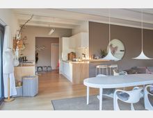 Offener Wohn- und Küchenbereich mit modernen Möbeln und hellen Farben. Die Küche besteht aus weißen Schränken und einer Holzarbeitsplatte; der Wohnbereich ist mit einem Esstisch, Designerstühlen und dekorativen Hängeleuchten ausgestattet.