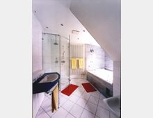 Modernes Badezimmer unter einer Dachschräge mit weißen Fliesen, einer gläsernen Duschkabine, einer Eckbadewanne mit blauen Verkleidungsfliesen, einem Waschbecken mit gelbem Handtuch, roten Teppichen und an der Wand aufgehängten gelben Handtüchern.