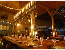 Langer, rustikaler Esstisch in einer Scheune mit Holzbalkendecke, dekoriert mit Kerzenleuchtern, Gläsern, Tellern und Blumenarrangements. Über dem Tisch hängt ein Kronleuchter aus Holz mit Kerzen sowie hängenden Blumen; im Hintergrund sichtbare Backsteinwände und dezente Beleuchtung.