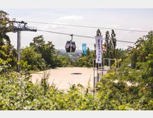 Eine Seilbahn-Gondel schwebt über einem hellen Platz, umgeben von Bäumen und Vegetation. Im Vordergrund sind Fahnenmasten mit mehrfarbigen Fahnen zu sehen, und im Hintergrund erstreckt sich eine weite Landschaft.