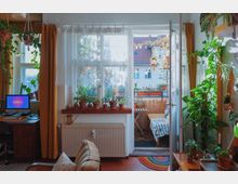 Wohnzimmer mit moderner Einrichtung, vielen Pflanzen in Töpfen und einem kleinen Arbeitsplatz mit Computer. Große Fenster öffnen sich zu einem Balkon mit Sitzmöbeln, Holzboden und weiteren Pflanzen; im Hintergrund ist ein gegenüberliegendes Wohngebäude sichtbar.