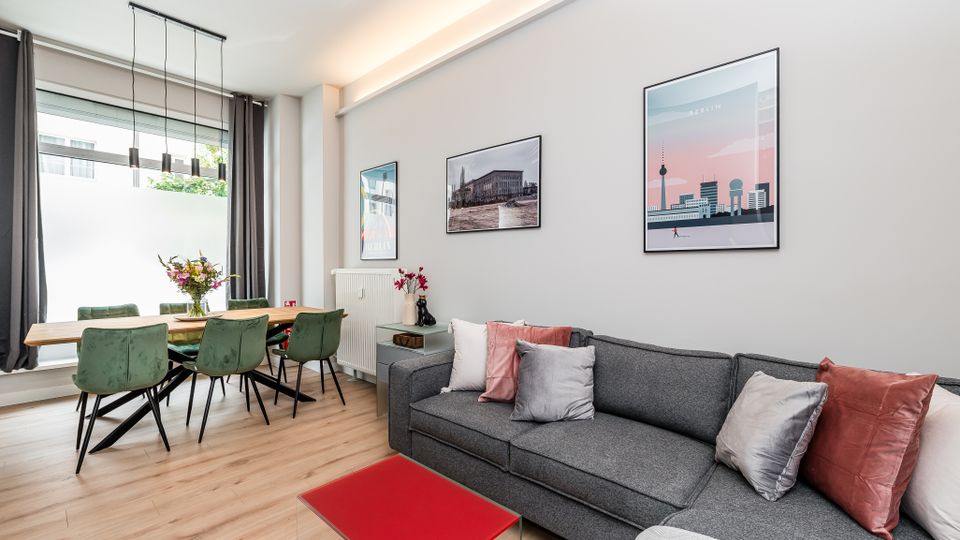 Heller, modern eingerichteter Wohn- und Essbereich mit großem Fenster und grauen Vorhängen, Holzboden sowie Esstisch mit grünen Stühlen und hängender Leuchte. Im Vordergrund steht ein graues Sofa mit Kissen und ein roter Couchtisch; an der Wand hängen gerahmte Bilder.