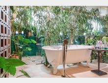 Ein Badezimmer mit freistehender weißer Badewanne auf Klauenfüßen, umgeben von zahlreichen großen grünen Pflanzen. Die Wände sind mit glänzenden, grünen Fliesen verkleidet, und ein Fenster lässt natürliches Licht hereinfallen.