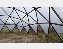 Ein verlassenes Geodät-Kuppelgewächshaus mit dreieckigen, durchscheinenden Kunststoffpaneelen auf einem Holzrahmen. Im Inneren ist der Boden von Gras und Unkraut bewachsen; im Hintergrund sind Bäume und Gebäude sichtbar.