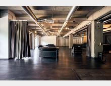 Großzügiger, offener Innenraum im Industriestil mit unverkleideter Betondecke, sichtbaren Lüftungsrohren und minimalistischer Beleuchtung. Der Raum ist mit schwarzen Ledersesseln, dunklem Bodenbelag und Vorhängen an den Seiten gestaltet.