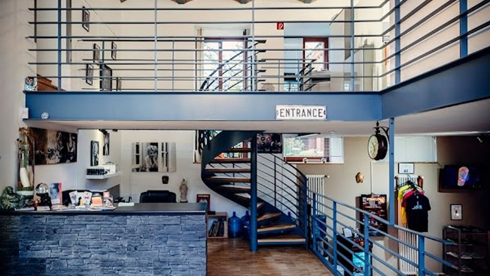 Großzügige Lobby mit zweigeschossiger Galerie, blauem Metallgeländer und zentraler Wendeltreppe; darüber eine hohe Ziegeldecke mit großem Kronleuchter. Im Vordergrund stehen eine Empfangstheke aus grauem Stein und ein Holzparkettboden, seitlich befinden sich Türen und Wanddekorationen.