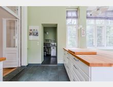 Helle, moderne Küche mit großer Kücheninsel und Arbeitsplatte aus Holz sowie weißen Schubladenschränken auf dunklem Fliesenboden. Große Sprossenfenster lassen viel Tageslicht herein; daneben befindet sich eine Nische mit kleinem Spülbereich.