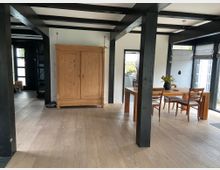 Wohnraum mit Holzboden und offenen schwarzen Deckenbalken. Im Vordergrund ein großer Holzschrank, daneben ein Esstisch mit Stühlen und einer Vase mit Tulpen; im Hintergrund sind große Fenster und ein Durchgang zur Küche sichtbar.