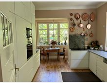 Helle Wohnküche mit weißen Einbauschränken, Holzfußboden und einer U-förmigen Arbeitsfläche mit Marmor-Rückwand. Vor großen Sprossenfenstern steht ein kleiner Holztisch mit zwei Stühlen, an der Wand hängen Kupfertöpfe und -pfannen.
