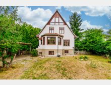 Zweistöckiges Wohnhaus mit Satteldach und verputzter weißer Fassade, umgeben von einem Garten mit Wiese, Bäumen und einem Holzzaun. Der Eingangsbereich auf der linken Seite ist teilweise von einem Holzvordach überdacht.