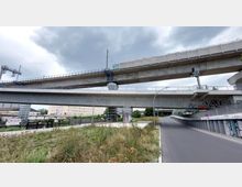 Eine urbane Szene mit zwei Betonbrücken, von denen die obere eine Bahntrasse trägt. Darunter verläuft eine Straße mit einer Abfahrt, umgeben von Grünflächen und Gebäuden im Hintergrund. Graffiti ist auf den Brücken und Wänden sichtbar.