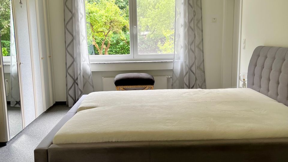 Helles Schlafzimmer mit großem Polsterbett und gepolstertem Kopfteil, grauem Teppichboden und einem Kleiderschrank mit Spiegeltüren. Ein breites Fenster mit hellen Vorhängen und Blick ins Grüne dominiert die Rückwand, darüber hängt eine Deckenleuchte.