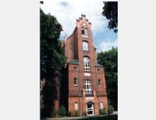 Backsteingebäude mit vier Stockwerken und einer Fassadengestaltung im Stil der Neogotik, erkennbar an Spitzbogenfenstern und Zinnen. Umgeben von grünen Bäumen, die das Gebäude teilweise einrahmen; der Himmel ist leicht bewölkt.