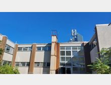 Flaches, funktionales Gebäude mit beigen Fassaden und großen Fenstern, teils verrosteten Metallstreben und einem Mobilfunkmast auf dem Dach; im Vordergrund vereinzelt grüne Pflanzen, blauer Himmel im Hintergrund.