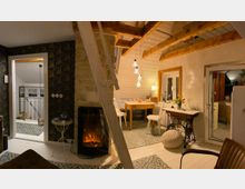 Gemütlicher Wohnraum mit Holzbalkendecke, Kamin, einer kleinen Sitzecke mit Holztisch und weißen Stühlen sowie dekorativem Teppichboden. Ein helles Fenster und eine Glastür bieten Ausblick, im Hintergrund ist eine offene Treppe erkennbar.