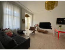 Wohnzimmer mit hellem Holzboden, großer schwarzer Couch mit Kissen, einem runden weißen Couchtisch und einem Sessel. An der Wand hängt ein Flachbildfernseher über einem schmalen TV-Unterschrank, daneben ein Regal. Im Hintergrund ein Esstisch mit weißen Stühlen und einer Tischplatte mit rot-orangefarbenen Beinen. Große Fenster mit weißen Vorhängen lassen Tageslicht herein.