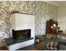 Wohnzimmer mit einem modernen Kamin mit dunkler Steinverkleidung, einer Tapete mit floralem Muster in Schwarz-Weiß und einem antiken Holzklavier mit Noten darauf. Im Raum befinden sich Tierkörbchen, ein Glas-Couchtisch und ein Sofa mit einer Felldecke.