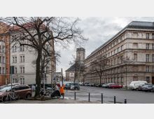 Straßenszene in einer Stadt mit mehrstöckigen, historischen Gebäuden aus Stein, teilweise mit Zierelementen versehen. In der Mitte ist ein Kirchturm mit Ziffernblatt sichtbar, im Vordergrund gepflasterte Bürgersteige, parkende Autos, Fahrräder und wenige Passanten.