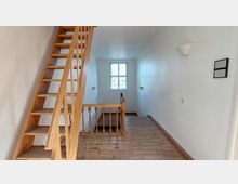 Flur mit Holztreppe, Holzboden und weiß gestrichenen Wänden. Am Ende des Flurs befindet sich ein Fenster mit Kreuzsprossen, das Tageslicht hereinlässt.