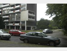 Mehrstöckiges Wohngebäude mit Balkonen und einer dunklen Fassade neben einer Baumreihe. Im Vordergrund stehen mehrere geparkte Autos auf einer gepflasterten Straße.