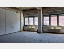 Leerer Raum in einem Gebäude im Rohbau mit unverputzten Wänden, drei großen Fenstern, Heizkörpern unter den Fenstern und einem Fliesenboden. Im Raum befinden sich eine Feuerlöscherflasche und eine Plastikflasche auf dem Boden.