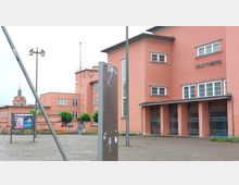 Außenansicht eines roten Gebäudes im Bauhausstil mit der Aufschrift „Stadttheater“ und Glasfenstern. Im Vordergrund befinden sich ein Infostand, Werbeplakate und eine gepflasterte Fläche, im Hintergrund ist ein Kirchturm sichtbar.