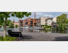 Holzpromenade mit einer Bank im Vordergrund, Blick auf eine Flusslandschaft mit Booten am Ufer und einer Reihe von Backsteingebäuden sowie modernen Wohnhäusern im Hintergrund. Eine Kirche mit einem spitzen Turm sowie üppige Bäume und eine Weide rahmen die Szene ein.