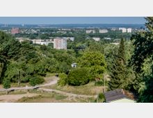 Blick auf eine grüne, leicht hügelige Landschaft mit Bäumen, Sträuchern und vereinzelten Gebäuden, im Hintergrund moderne Wohnblöcke und eine größere Backsteinkirche. Der Vordergrund zeigt einen unbefestigten Weg und eine kleine Hütte mit flachem Dach.