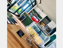 Innenansicht eines modernen Cafés mit einem Tresen, der mit grünen Fliesen verkleidet ist. Im Vordergrund steht ein gedeckter Kuchenbehälter mit Zitronenkuchen auf einem Holztisch, daneben ein kleines Schild mit der Preisinformation; im Hintergrund sind Regale mit Getränken, ein roter Kühlschrank und große Fenster zu sehen.