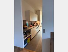 Blick in eine moderne, schmale Küche mit hellen Einbauschränken, einer grauen Arbeitsplatte und grauen Fliesen auf dem Boden. Links sind offene Regale mit Küchenutensilien, rechts ein Herd mit darüberliegenden Hängeschränken; der Raum ist gut beleuchtet durch Tageslicht.