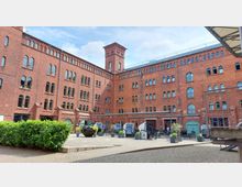 Roter Backsteinbau mit mehreren Stockwerken und vielen Rundbogenfenstern, im Innenhof befinden sich Sitzgelegenheiten mit Sonnenschutz und Pflanzen in Töpfen. Im Hintergrund sind Eingänge mit Schildern zu einem "Brauhaus" und "Speicher" sichtbar.