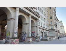 Straßenszene mit einem großen, hellen Steinbau im klassizistischen Stil, dessen Erdgeschoss aus hohen Rundbögen und massiven Säulen besteht und mit Graffiti bedeckt ist. Entlang der Fassade verläuft ein breiter Gehweg; im Hintergrund ist ein hoher Turm mit grüner Kuppel zu sehen.