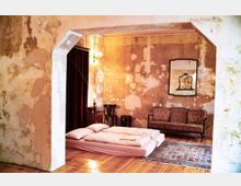 Schlafbereich in einem Raum mit abgenutzten Wänden, einem Holzboden, zwei gestapelten Matratzen mit Bettzeug auf einem Teppich sowie einem antiken Sofa und einem Spiegel an der Wand. Im Hintergrund sind eine rote Samtvorhänge und eine kleine Lampe sichtbar.