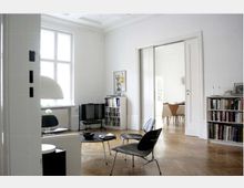 Wohnzimmer in einem hellen, modernen Apartment mit weißen Wänden, hohen Decken und Holzparkettboden. Der Raum ist mit minimalistischen Möbeln eingerichtet, darunter schwarze Stühle, ein kleiner Couchtisch und ein Regal mit Büchern; im Hintergrund führt ein offener Durchgang in einen angrenzenden Essbereich.