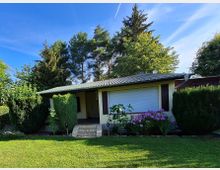 Kleines Einfamilienhaus mit Flachdach und heller Fassade, umgeben von einer gepflegten Rasenfläche, blühenden Blumenbeeten, Büschen und hohen Bäumen im Hintergrund. Der Eingang ist über eine kurze Treppe mit Ziegelstufen erreichbar.