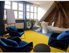 Heller Besprechungsraum mit modernem Design, ausgestattet mit blauen Sesseln, einem runden Holztisch, weißen Stühlen und einer gelb-blauen Bodenfläche. Im Hintergrund sind große Fenster, eine Backsteinwand mit freiliegenden Holzträgern sowie ein Flipchart und Zimmerpflanzen zu sehen.