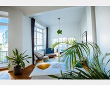 Helles, modernes Zimmer mit großen Fenstern, Blick auf ein Außenbalkon und städtische Gebäude. Im Raum befinden sich ein Bett mit gelbem Kissen, Zimmerpflanzen, ein kleiner Fernseher und minimalistische Möbel.