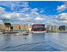 Moderne Architektur am Ufer eines Flusses, darunter ein braunes Gebäude mit kantigen Formen und einem überhängenden Glasaufsatz, sowie weitere Gebäude mit Glasfassaden und Balkonen. Im Vordergrund fährt ein weißes Boot auf dem Wasser, während der Himmel teilweise bewölkt ist.