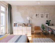 Minimalistisch eingerichtetes Zimmer mit Holzboden, einer verputzten Wand und großen Fenstern auf der linken Seite. Zu sehen sind ein Regal mit Büchern, ein Stuhl, Pflanzen, ein Schreibtisch und drei Bilder an der Wand.