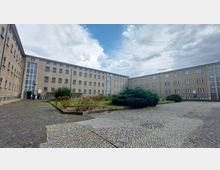 Hof eines historischen, rechteckigen Gebäudekomplexes mit mehreren Stockwerken aus grauem Putz, rechteckigen Fenstern und Glaselementen. In der Mitte des gepflasterten Hofes befinden sich Grünflächen mit Blumenbeeten und Büschen.