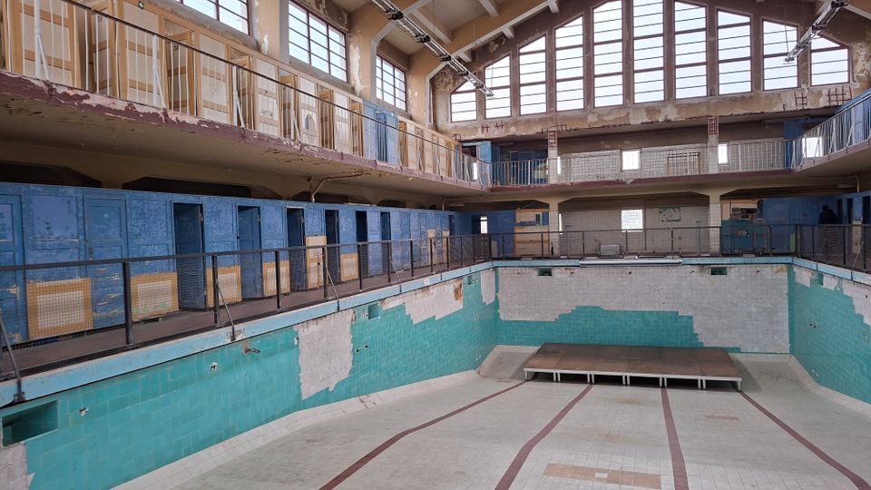 Ehemaliges Hallenbad mit leerem, gefliestem Becken in einem verfallenen Zustand. Die Wände sind teilweise mit blauen und türkisfarbenen Fliesen bedeckt, im oberen Bereich befinden sich alte Umkleidekabinen und eine umlaufende Galerie mit Geländern, darüber große Fensterflächen.