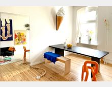 Heller Raum mit Holzfußboden, einem schwarzen Tisch, Holzbänken und einem orangen Stuhl aus geschwungenem Material. An der weißen Wand hängen kunstvolle Gemälde und eine Wanddekoration in Form eines Eiswaffelhorns; daneben ein großes Fenster mit weißen Vorhängen und Blumen auf der Fensterbank.