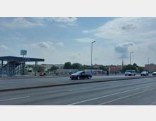 Straßenszene in der Nähe des S-Bahnhofs Westhafen in Berlin, mit einer erhöhten Bahnzugangsanlage, Fahrradständern und mehreren parkenden Fahrrädern im Vordergrund. Im Hintergrund sind Wohnhäuser, Bäume und ein Kirchturm zu sehen, während Autos und Fußgänger die Straße überqueren.