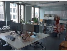 Modernes Großraumbüro mit mehreren Arbeitsplätzen, ausgestattet mit Schreibtischen, Monitoren, Kabeln und Bürostühlen. Große Fenster lassen Tageslicht herein, und Pflanzen sowie Regale mit Büromaterialien ergänzen die Einrichtung.