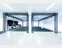 Ein großer, heller Raum mit schwarzen Metallrahmen-Strukturen, weißen Wänden und dunklem Bodenbelag. Im Hintergrund befinden sich Arbeitsstationen mit Computern und technischen Geräten.